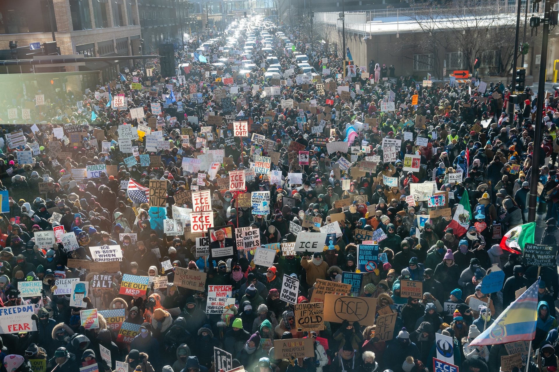 A large aerial view of a dense urban protest with thousands of people holding varied signs opposing ICE and immigration enforcement, filling a city street with buildings on either side.